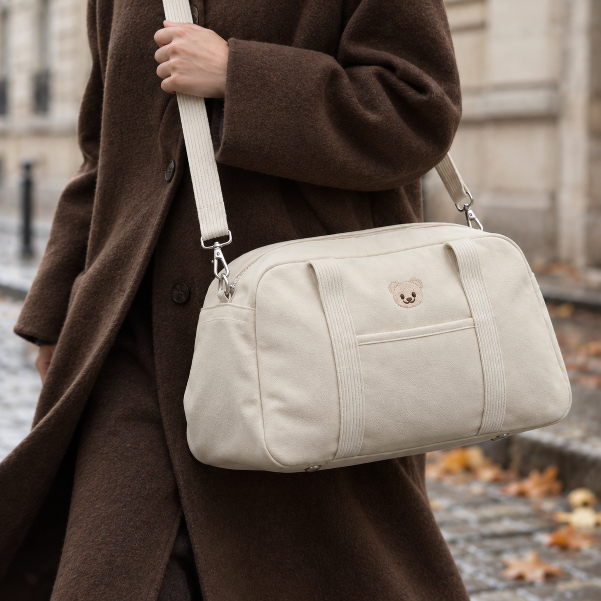 Sac à langer week-end NEVA Teddy porté en bandoulière sur un manteau camel, dans une rue parisienne en automne