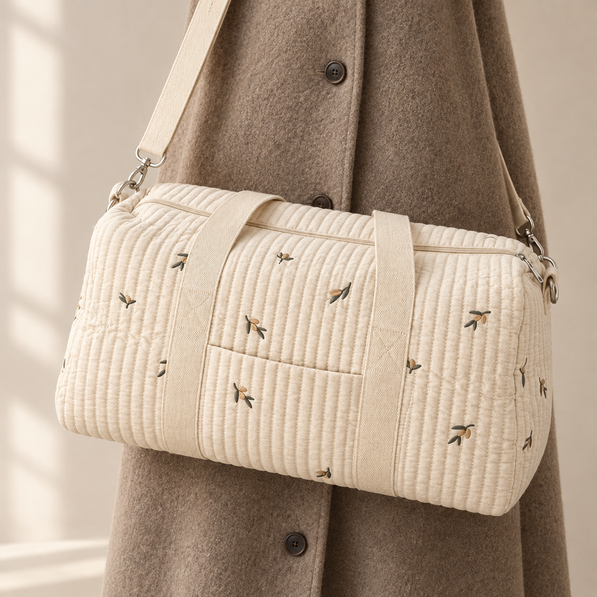 Sac à langer L’Intégral brodé olives tenu à la main par ses anses, dans une entrée parisienne au parquet en chevrons