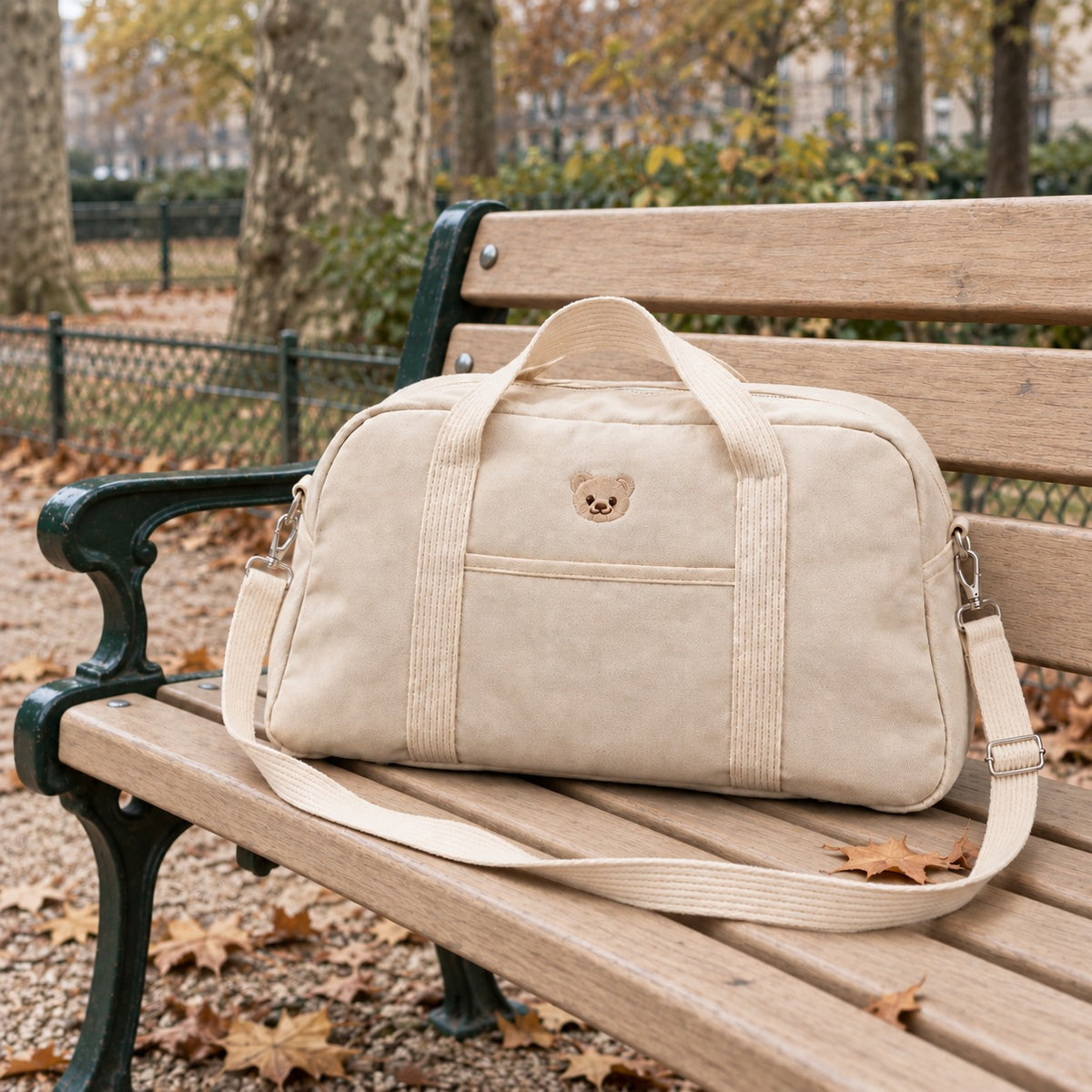 Sac à langer week-end NEVA Teddy en toile crème posé sur un banc de jardin public parisien